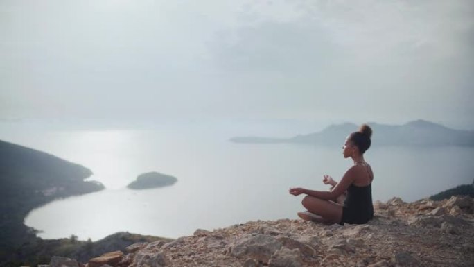 非洲族裔妇女在悬崖顶上冥想，看着莲花位置的阳光明媚的大海