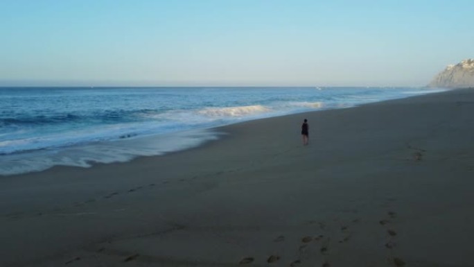 空中无人机拍摄了一名妇女在日出时沿着沿海度假胜地海滩行走的镜头