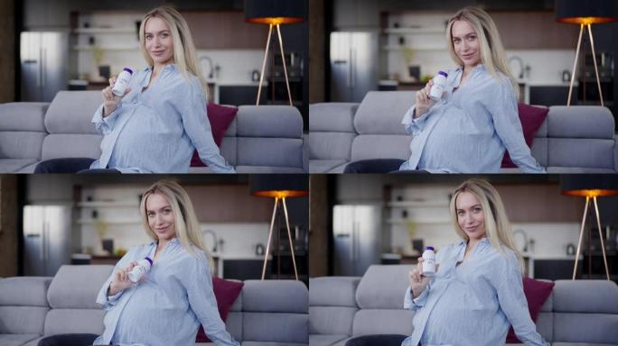 微笑自信的孕妇拿着维生素D3瓶看着坐在客厅沙发上的相机。美丽的高加索人期待在家中室内广告健康维生素的