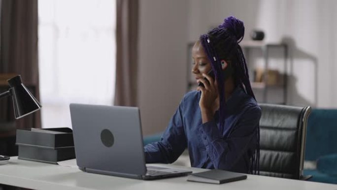 年轻开朗的黑人女商人在她的家庭办公室里用手机聊天，坐在笔记本电脑的桌子旁，用手机聊天