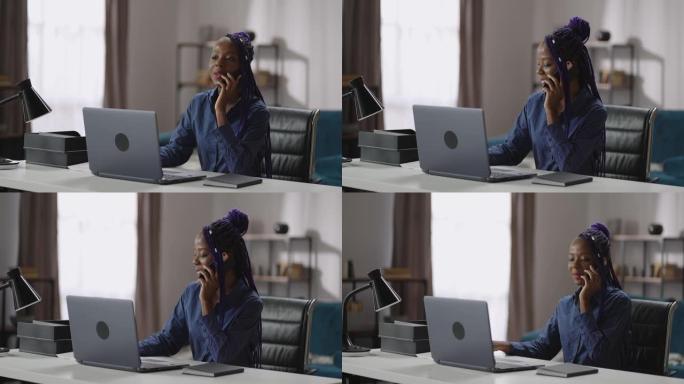 年轻开朗的黑人女商人在她的家庭办公室里用手机聊天，坐在笔记本电脑的桌子旁，用手机聊天