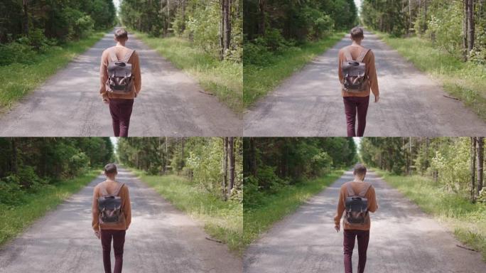 一个背着背包的年轻人走在树林里的一条土路上，看着地形。后视图。