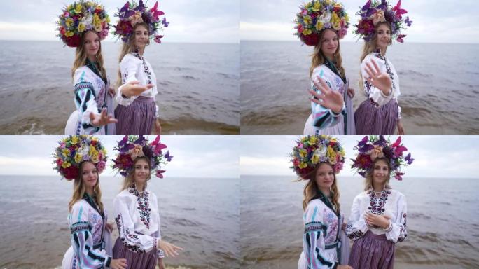 两名穿着传统花圈和民族服装的年轻乌克兰妇女站在河边海滩上慢动作地向镜头伸展双手。自信的女士在户外摆姿