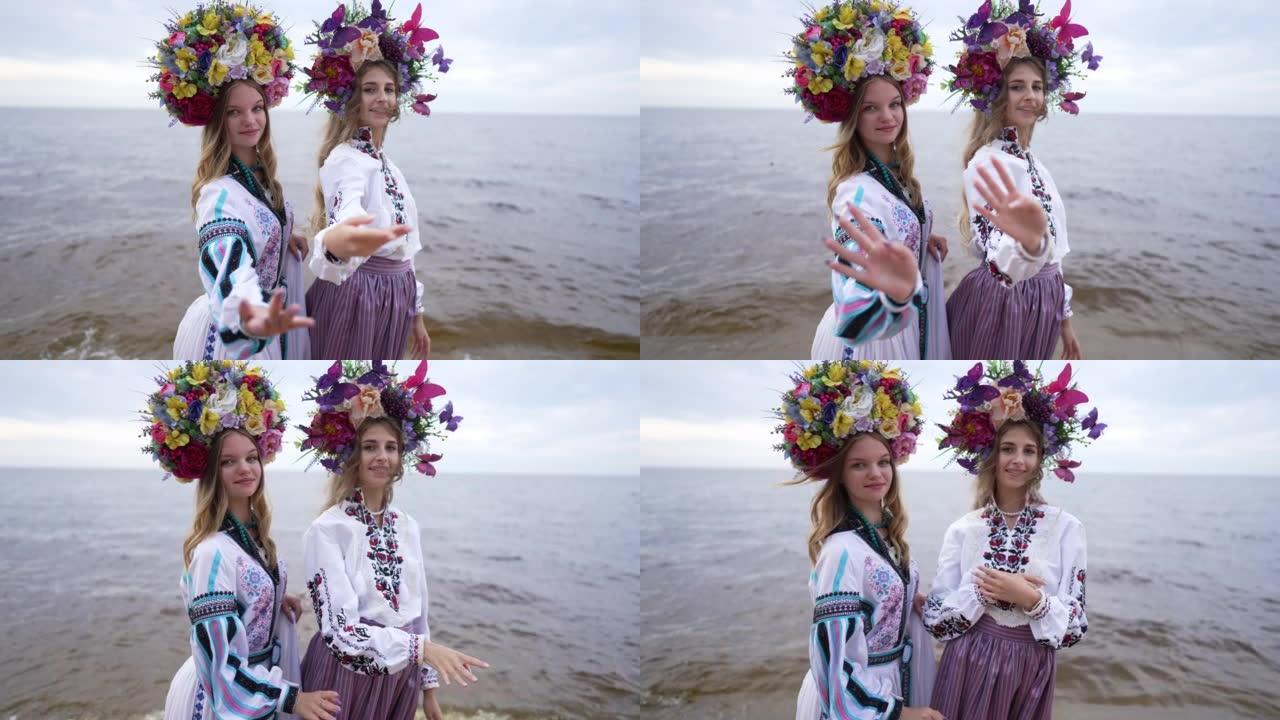 两名穿着传统花圈和民族服装的年轻乌克兰妇女站在河边海滩上慢动作地向镜头伸展双手。自信的女士在户外摆姿