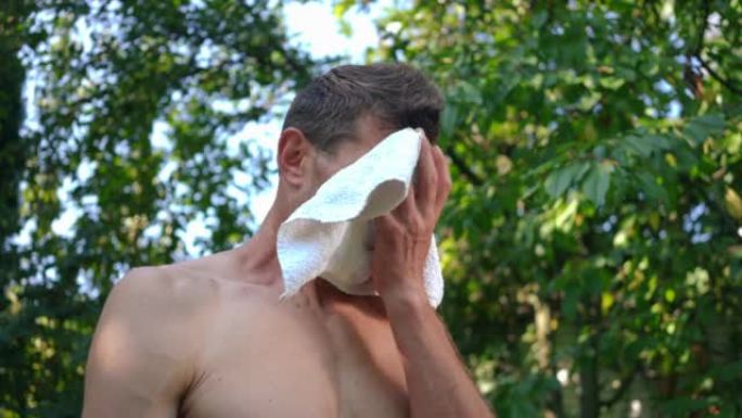 出汗合身的运动员在夏天站在户外用白毛巾擦脸。在后院训练后疲惫的高加索人的肖像。健康的生活方式和力量理