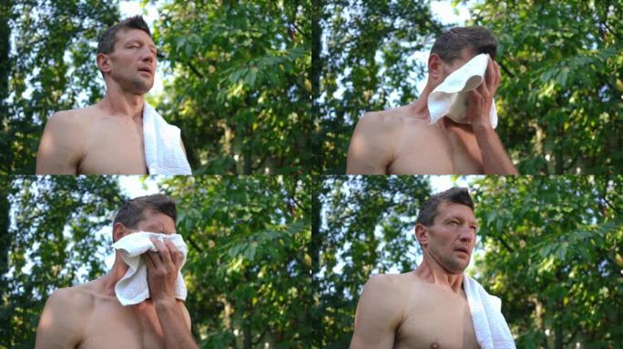 出汗合身的运动员在夏天站在户外用白毛巾擦脸。在后院训练后疲惫的高加索人的肖像。健康的生活方式和力量理