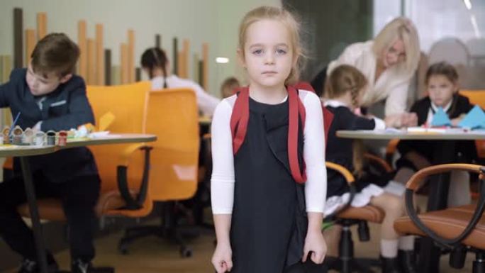 极其美丽的小女孩背着背包站在教室里，背景模糊的同学和老师。可爱的积极的高加索女学生在镜头前摆姿势微笑