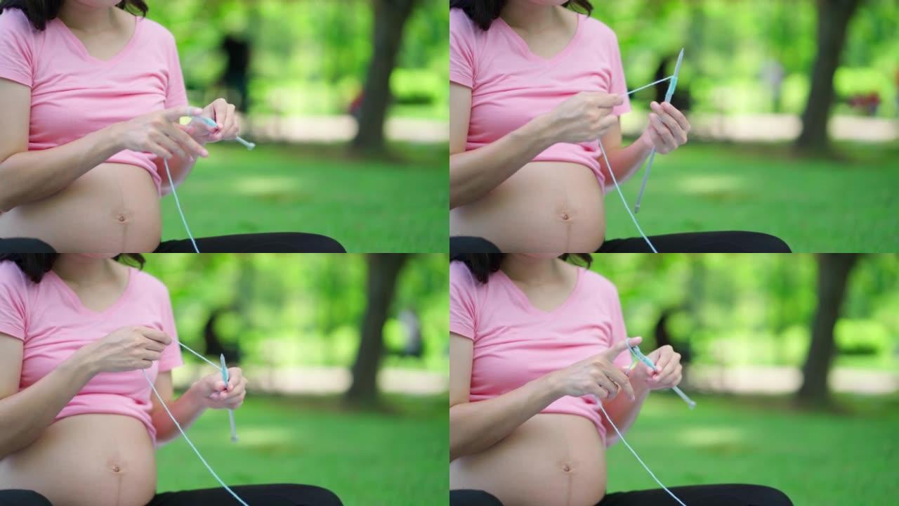 漂亮的亚洲孕妇微笑着在公共花园里为婴儿织针。户外家居用蓝色织物手工编织工艺。为婴儿创造手工配件