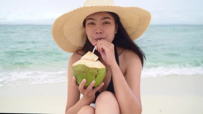 在晴朗的日子里，亚洲妇女坐在椅子上在海滩上喝椰子水