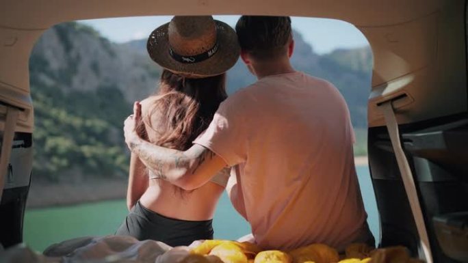 可爱的年轻夫妇坐在美丽的绿松石湖边的汽车后备箱里拥抱