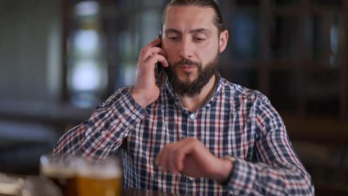 英俊的男人坐在室内的酒吧柜台接电话聊天。周末在酒吧里等着朋友的自信的大胡子高加索人的肖像。休闲和生活