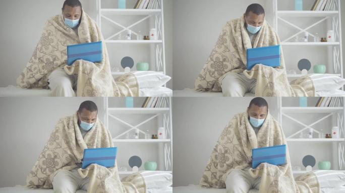 戴着口罩的颤抖的男人坐在医院病房里，使用笔记本电脑。生病的非洲裔美国人上网的肖像。有冠状病毒病毒性疾