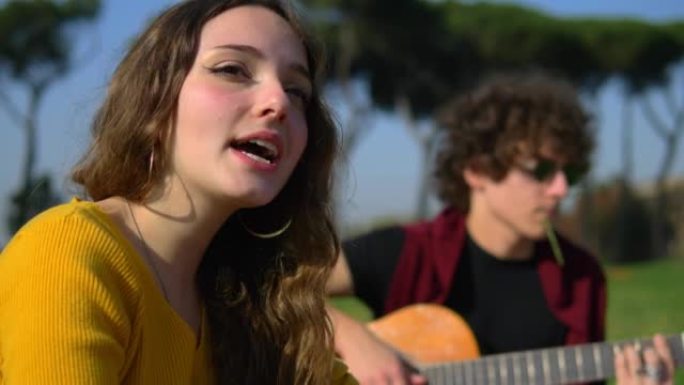 坐在草坪上的年轻无忧无虑的女人和弹吉他的朋友一起唱歌