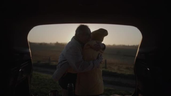 两个老年人在公路旅行中露营。站在大型汽车后备箱旁拥抱起来