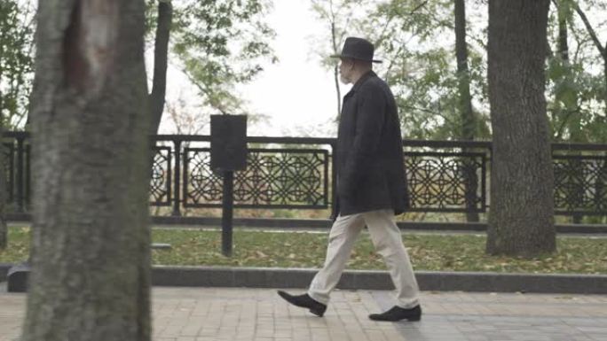 在阴天，相机跟随穿着优雅服装的谨慎间谍或侦探沿着公园小巷行走。穿着帽子和外套的自信的白人男子在户外漫