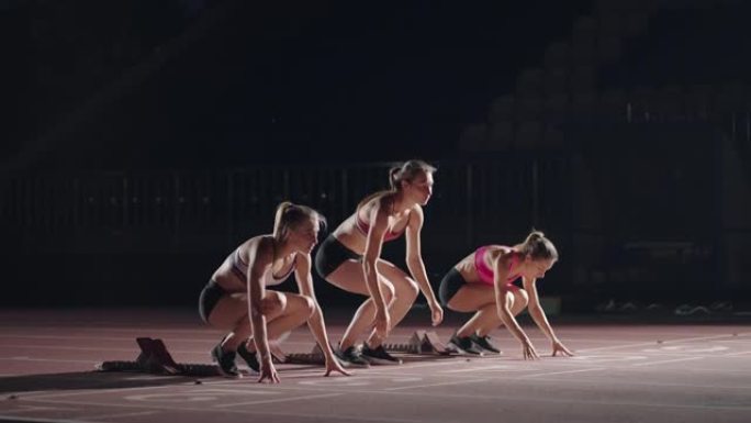 一排跑步者女子在开始比赛前蹲在起跑位置。雌性从体育场的起跑鞋开始，在黑暗中，聚光灯在慢动作中。