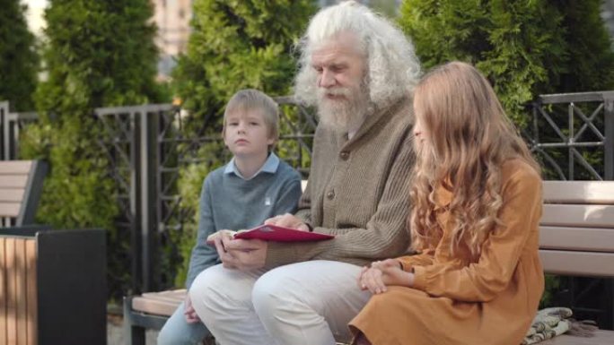 聪明的男性退休人员在户外晴天向孙子解释书的角度视图。自信快乐的高加索祖父为坐在长凳上的男孩和女孩读书