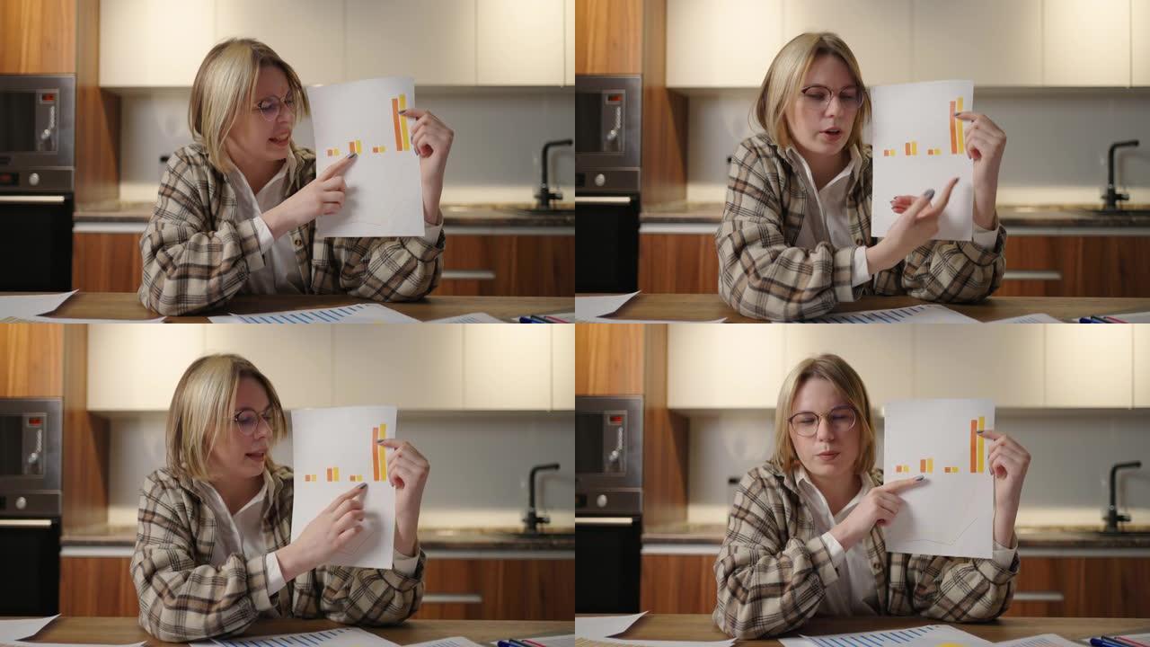 看着家里戴眼镜的女人坐在厨房里，对着网络摄像机显示一张图表。家庭办公室远程工作。用图表和表格进行视频