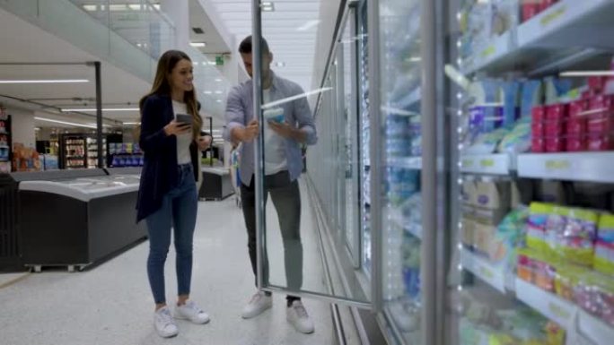 快乐的拉丁美洲夫妇在冷藏区的超市购买乳制品，并在智能手机上跟踪购物清单