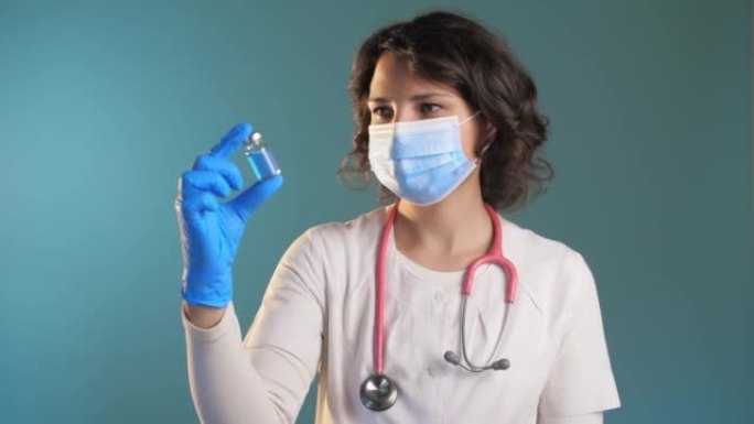 戴着医用口罩的女医生站在蓝色背景下，看着安瓿的疫苗。蓝色药瓶。慢动作