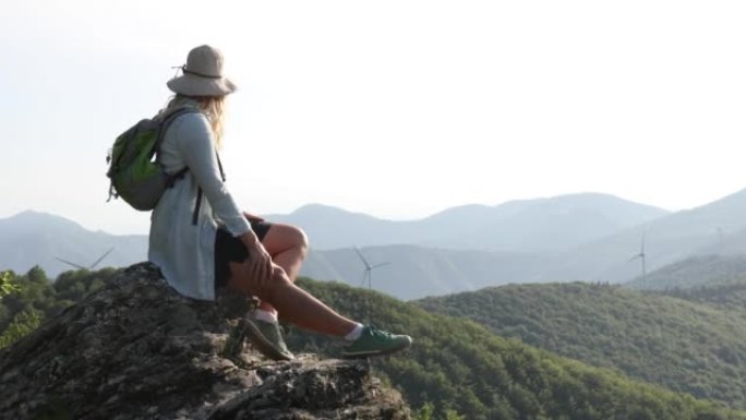 女徒步旅行者到达绿山风力涡轮机上方的岩石山顶