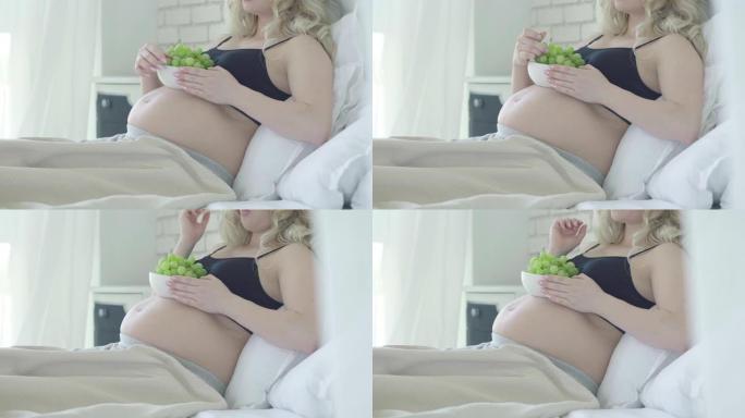无法辨认的孕妇躺在床上，吃健康的葡萄。放松的白人女士在卧室里在家享受健康的食物。快乐怀孕概念。