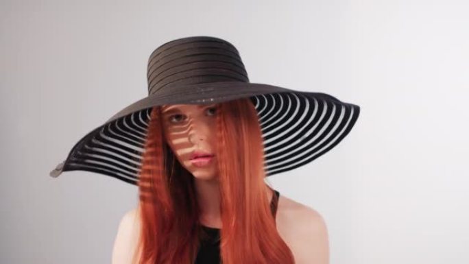 美丽诱人的红发女人用相机调情。戴着大而优雅的帽子摆姿势，遮住她的脸