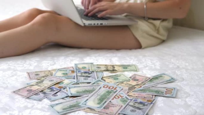 一个女孩坐在床垫上的特写镜头，在零散的美元货币的背景下在笔记本电脑上打印。自由职业者在家赚钱，赚钱。