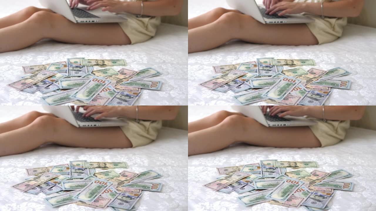 一个女孩坐在床垫上的特写镜头，在零散的美元货币的背景下在笔记本电脑上打印。自由职业者在家赚钱，赚钱。