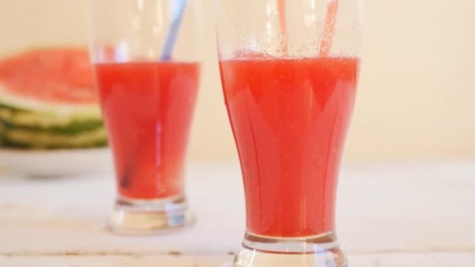特写两个玻璃玻璃杯，用西瓜果肉中的红色鲜榨饮料站在白色桌子上，靠近一个躺着的成熟的红色西瓜，并带有可