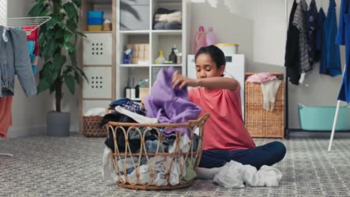 微笑的女孩坐在洗衣房地板上，在大柳条篮前，衣服要分类，将空闲时间花在家务上，帮助父母