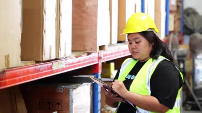 亚洲肥胖的女性仓库工人在仓库工厂工业中穿着安全背心和安全帽头盔。货架上纸箱中的产品。工作时加大码女性