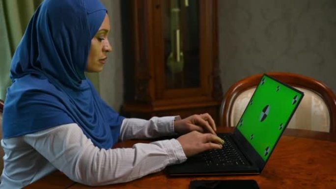 戴着头巾的勤奋的穆斯林妇女，在带有绿色色度键的笔记本电脑上远程工作。广告剪辑的模型屏幕