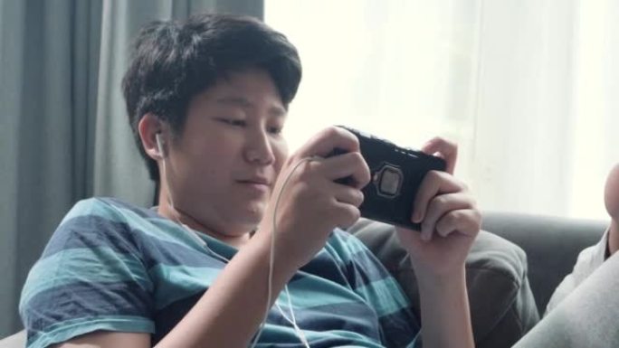 亚洲少年男孩躺在沙发上，在家窗口附近玩手机游戏，生活方式概念。