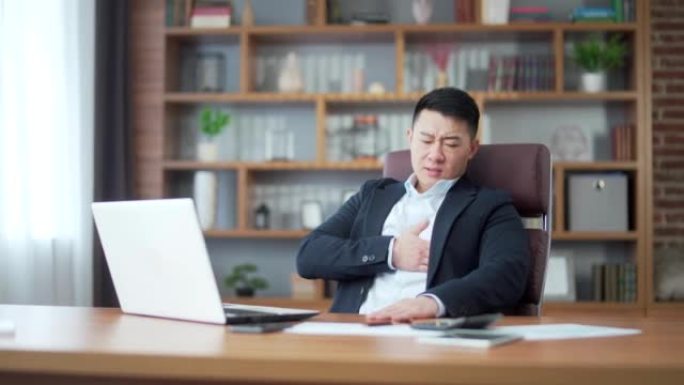 亚洲男性商人办公室职员，严重的心脏疼痛。胸口憋屈，心脏病发作。商务男士雇员坐在电脑桌前工作。在职场中