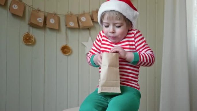 穿着睡衣的男孩，圣诞老人的帽子伸展着带礼物的纸质降临日历。小快乐好奇的孩子打开手工圣诞日历的礼物挂在
