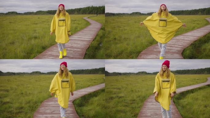 在多云的天气里，戴着红色帽子和黄色雨披雨衣的漂亮女人喜欢在森林沼泽中的木路上散步