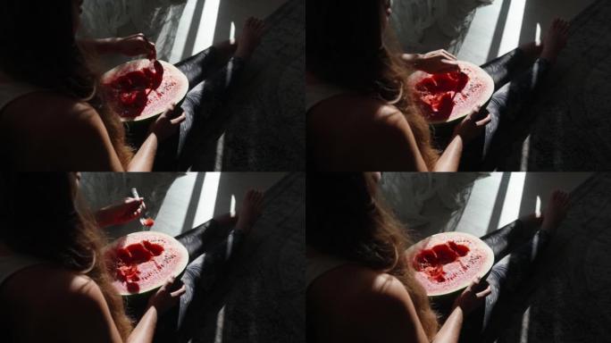 女孩坐在地板上，拿着一个大西瓜，她用勺子吃。阳光下的西瓜