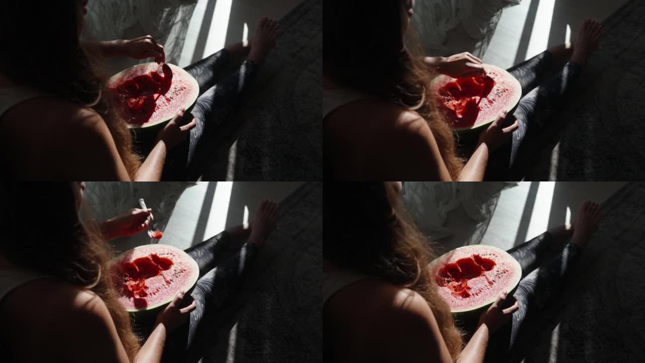 女孩坐在地板上，拿着一个大西瓜，她用勺子吃。阳光下的西瓜