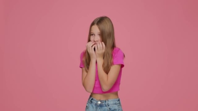 紧张害怕的小学生女孩在粉红色的工作室背景上用手指遮住嘴。恐慌，恐惧症，焦虑