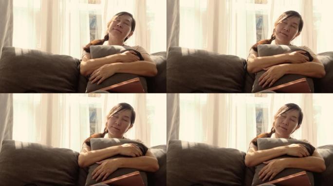 昏昏欲睡的亚洲成年女性坐在沙发上，靠着阳光在家靠近窗户，生活方式的概念。