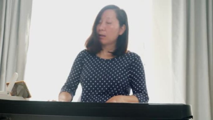 亚洲成年妈妈通过手机应用程序在家学习在窗户附近玩键盘乐器，生活方式概念。