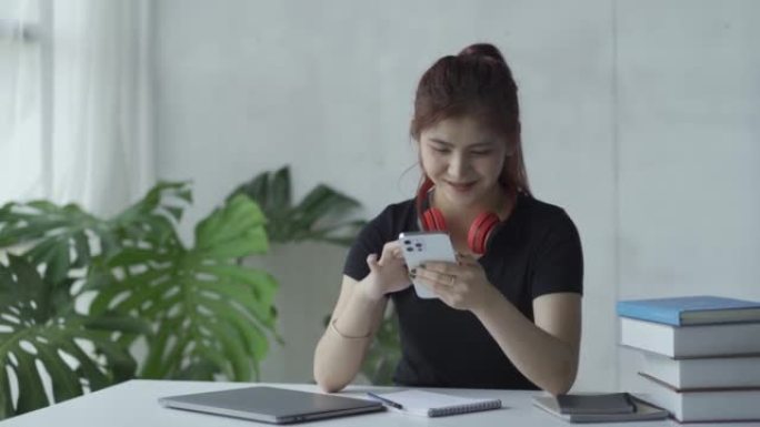 亚洲妇女戴着耳机在智能手机上聊天，笔记本电脑和虎皮在学习概念桌上聊天