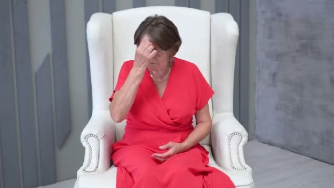 一位70岁的老妇人穿着红色连衣裙，戴着眼镜，坐在家里的扶手椅上思考和担心孩子，在胸前画十字。老年人及