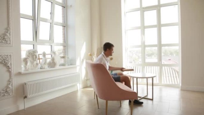 一个男人坐在宽敞房间的沙发上，从咖啡桌上拿一本书，开始阅读