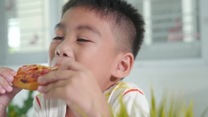 快乐的亚洲小男孩5年坐在家里的桌子上吃巨大的披萨