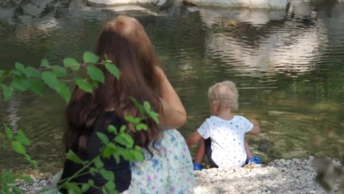 妈妈看着她的小孩在夏天公园的河岸上玩耍，1.5岁的小孩喜欢把鹅卵石扔进水里。