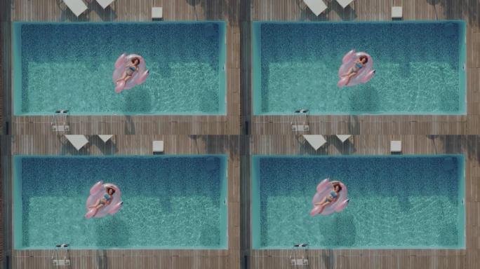在炎热的夏日，一位长发的年轻旅游妇女在游泳池中间的充气粉红色火烈鸟上晒日光浴。女性在阳光明媚的日子里