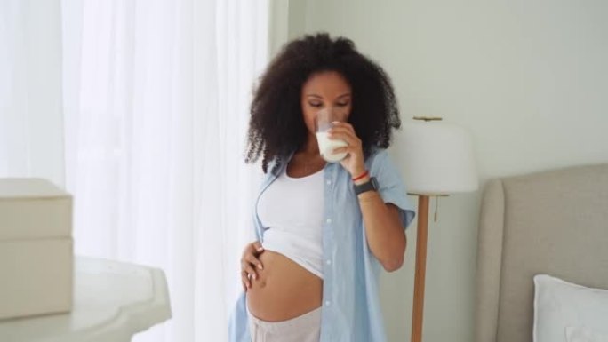 怀孕的年轻妇女在窗户附近穿着便服在家喝一杯牛奶。享受怀孕，怀孕时健康饮食的概念。乳制品营养的好处。
