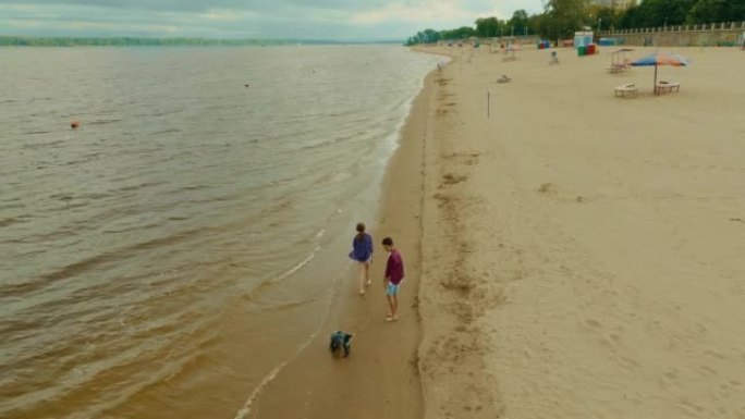 鸟瞰图，孩子们带着小狗在河岸上散步。男孩和一个女孩光着脚沿着河边散步，一只狗在他们旁边奔跑。美丽的风